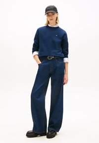 Kvinna som bär en marinblå sweatshirt över en vit skjorta, vida mörka jeans, svart bälte, svarta platåskor och en svart keps, stående med händerna i fickorna.