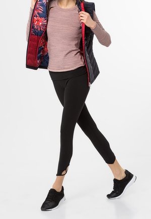 Schwarze Leggings mit Ausschnitten, hellrosa Langarmshirt und eine gepolsterte Weste mit floralem Innenfutter, kombiniert mit schwarzen Sportschuhen.