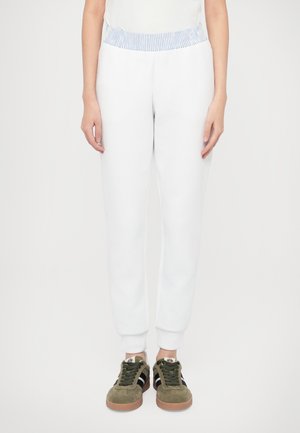 Pantaloni della tuta bianchi con una fascia elastica a righe azzurro chiaro, polsini aderenti, abbinati a sneakers verdi in suede con strisce nere.