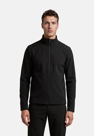 Hombre con una chaqueta negra con cremallera, cuello alto y mangas largas, de pie frente a un fondo liso gris claro.