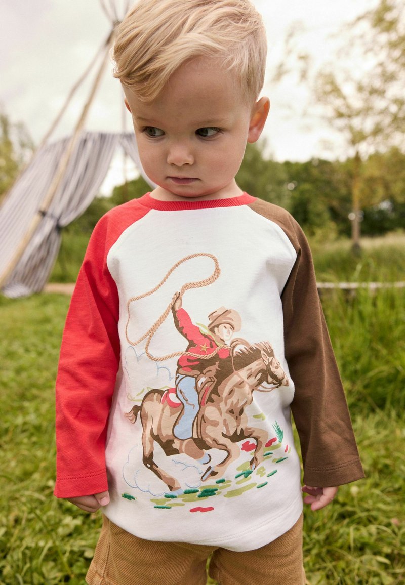 Ung blondt barn iført en rød og brun langærmet trøje med en grafik af en cowboy, der rider på en hest, stående udenfor på græs.
