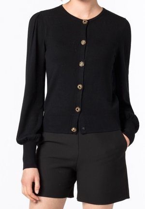 Vrouw draagt een zwart lange-mouwen vest met zes decoratieve gouden knoppen en zwarte shorts met hoge taille, met één hand in de zak.