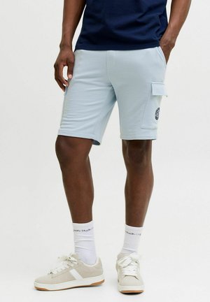 Mężczyzna ubrany w jasnoniebieskie krótkie spodenki typu cargo z naszywką, białe skarpetki do kostki i beżowe sneakersy na jednolitym tle.