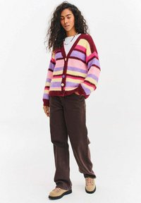 Kardigan wielokolorowy z poziomymi paskami w kolorze różowym, fioletowym, żółtym i czerwonym, z dekoltem w kształcie litery V, z przodu zapinany na guziki, wykonany z dzianiny o fakturze. Połączony z ciemnymi spodniami.