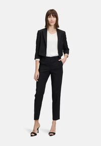 Completo nero su misura con giacca corta e pantaloni sartoriali, abbinato a una blusa plissettata bianca e tacchi neri. Texture liscia, design strutturato.