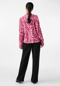 Long-sleeve blouse in pink with a floral pattern, featuring a gathered back, paired with black wide-leg trousers and black heels.