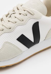 Adidași din plasă albă cu accente din daim. Prezintă logo-ul V negru, material texturat și talpă bej. Închidere cu șireturi, design elegant.