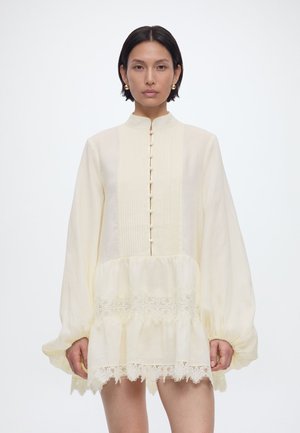 Kvinde med kort mørkt hår iført en cremet langærmet kjole med knapper, blonder detaljer og opsamlede manchetter mod en ensfarvet hvid baggrund.