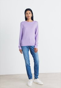 Sweater lilás de mangas longas e decote redondo, combinado com jeans azuis e ténis brancos, em frente a um fundo neutro claro.