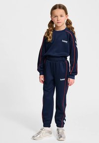 Marineblauer Trainingsanzug aus weichem Material, mit roten Streifen und weißem Logo. Enthält langärmliges Oberteil und Hosen mit elastischem Bund. Silberne Turnschuhe.