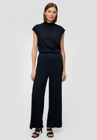Navy geribbelde jumpsuit met een hoge kraag en kapmouwen; heeft brede, vloeiende pijpen en een ontspannen pasvorm; gedragen met zwarte haksandalen.