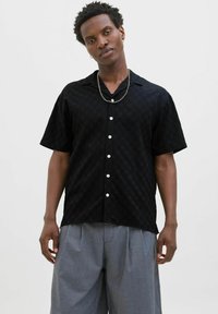 Chemise noire à manches courtes avec un motif texturé, fermeture à boutons et boutons blancs. Portée avec un pantalon gris à plis; accessoire en chaîne subtil.