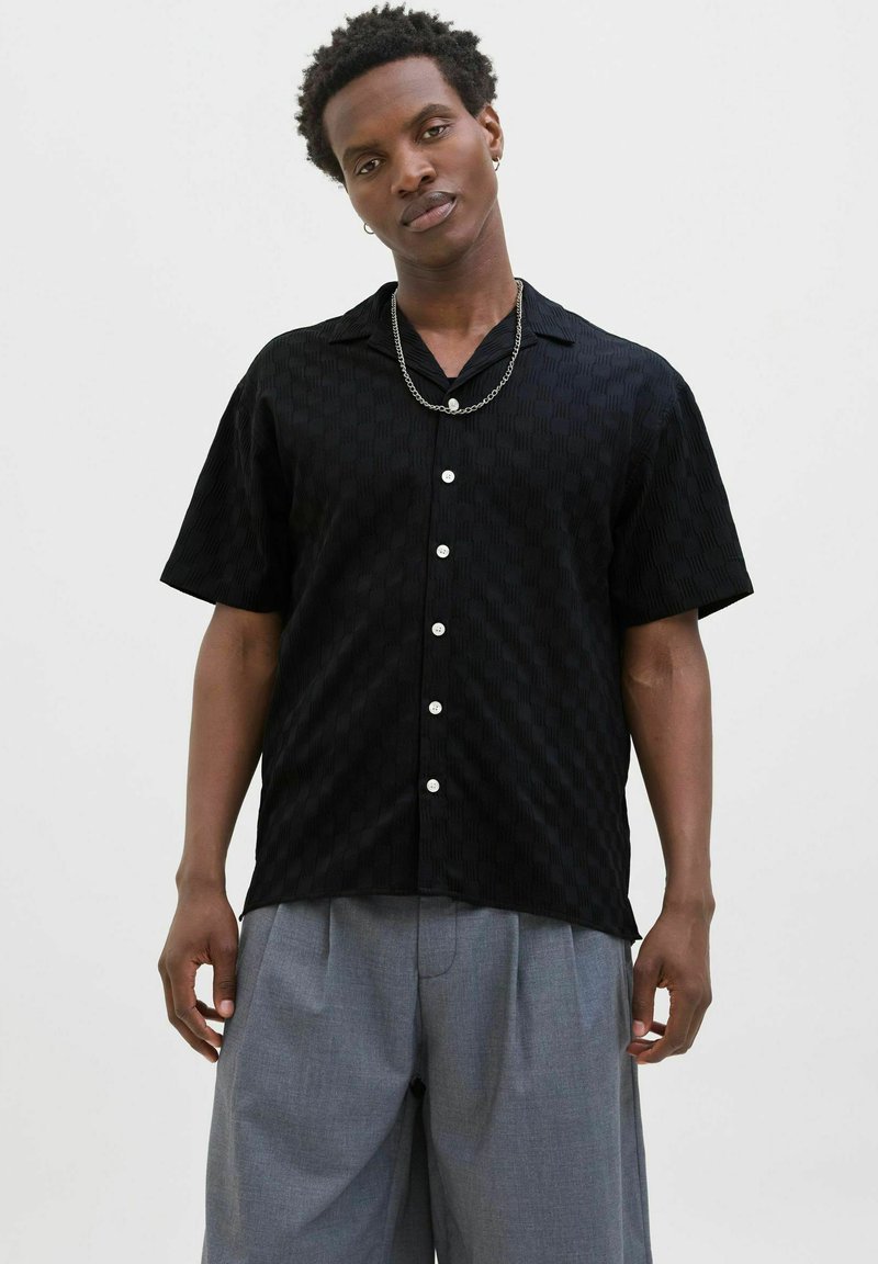 Chemise noire à manches courtes avec un motif texturé, fermeture à boutons et boutons blancs. Portée avec un pantalon gris à plis; accessoire en chaîne subtil.