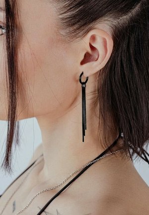 Seitenprofil einer Frau mit schwarzem langen Quastenohrring, dunklem Haar hinter dem Ohr zurückgesteckt und einer silbernen Kettenhalskette, die auf der Schulter sichtbar ist.