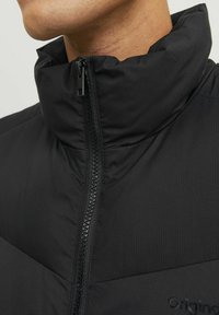 Schwarze gepolsterte Jacke mit hohem Kragen, ausgestattet mit einem Frontreißverschluss und strukturierten Stoffdetails. Logo an der unteren Seite platziert.