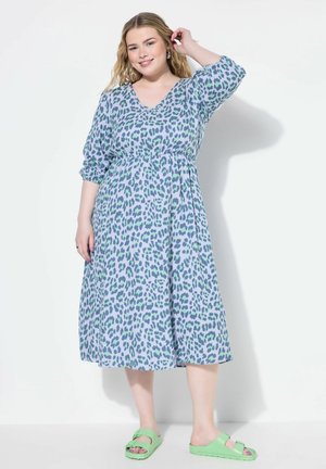 Leopardprint jurk van lichtblauwe stof, met een V-hals, geplooide taille en 3/4 mouwen. Gecombineerd met groene schuifsandalen.