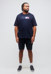 Άνδρας με κοντά dreadlocks που φοράει μπλε ναυτικό μπλουζάκι Tommy Hilfiger, ταιριαστό σορτς, λευκές κάλτσες και μαύρα αθλητικά παπούτσια, στέκεται μπροστά σε λευκό φόντο.