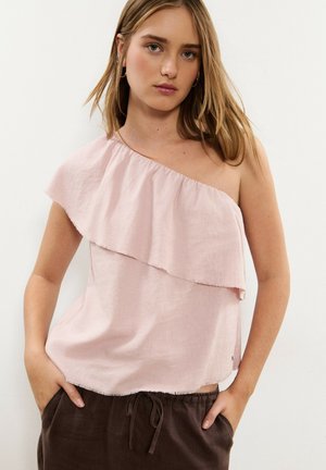 Mujer joven con un top de un solo hombro color rosa pálido con volantes y pantalones marrón oscuro, con las manos en los bolsillos contra un fondo liso.