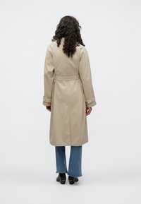 Trench-coat beige avec taille ceinturée, manches longues et détails aux poignets. Porté avec un jean évasé et des bottines noires à talons. Design minimaliste, texture lisse.