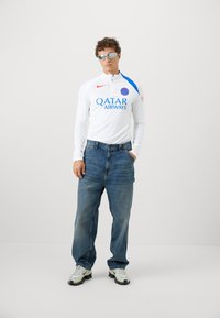 Hvid langærmet sportsjersey med røde og blå detaljer, der har "QATAR AIRWAYS" logoet. Parret med løse blå jeans og hvide sneakers.
