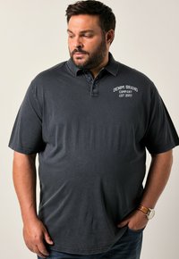Polo tričko v barvě uhlově šedé s límcem na dvě knoflíky, vyšitým logem na hrudi a krátkými rukávy. Nosené s tmavými džíny a hodinkami.