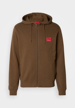 Brūns džemperis ar iešuvuma rāvējslēdzēju, ar kangarū atstarpēm, regulējamu kapuci ar auklu un sarkanu taisnstūrveida uzlīmi ar uzrakstu "HUGO" uz krūtīm.