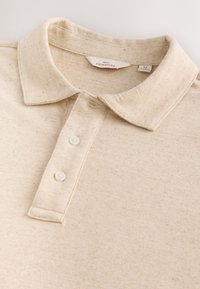 Tricou polo bej din material texturat, cu guler clasic, patru nasturi și model discret cu pete. Eticheta poartă inscripția "NEXT SIGNATURE".