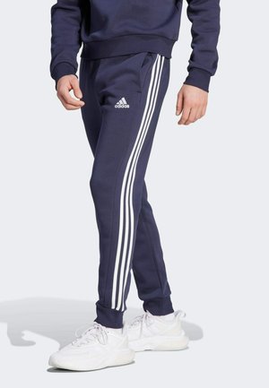Mørkeblå træningsbukser med tre hvide striber på siderne, elastiske manchetter og en Adidas-logo på venstre lår.