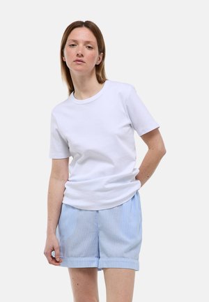 Femme portant un T-shirt blanc uni à manches courtes et un short rayé bleu clair, debout devant un fond blanc.