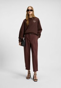 Felpa marrone con logo, pantaloni marroni a vita alta con cintura e tacchi neri con fasce. La modella indossa occhiali da sole neri e ha i capelli lisci.