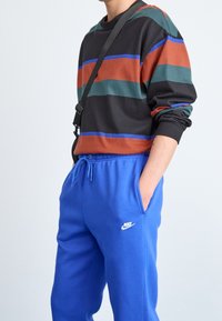 Multicolored long-sleeve shirt with horizontal stripes (black, green, rust, blue) paired with blue sweatpants featuring a white logo.
