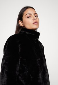 Manteau en fausse fourrure noire avec un col montant, texture douce et design épuré, présentant une silhouette ajustée et des détails de couture minimaux.