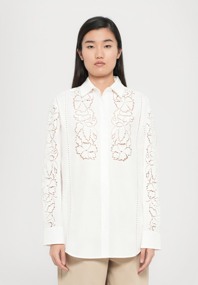 Camisa blanca de botones con bordado floral en las mangas, con cuello puntiagudo, mangas largas y un ajuste relajado. Textura de tela suave.