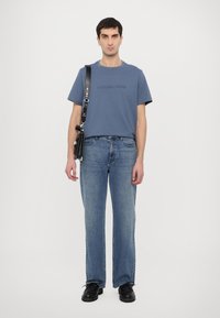 Kék pamut póló domború "MICHAEL KORS" logóval, világoskék farmernadrággal párosítva, és fekete válltáskával. A cipők feketék és masszívak.