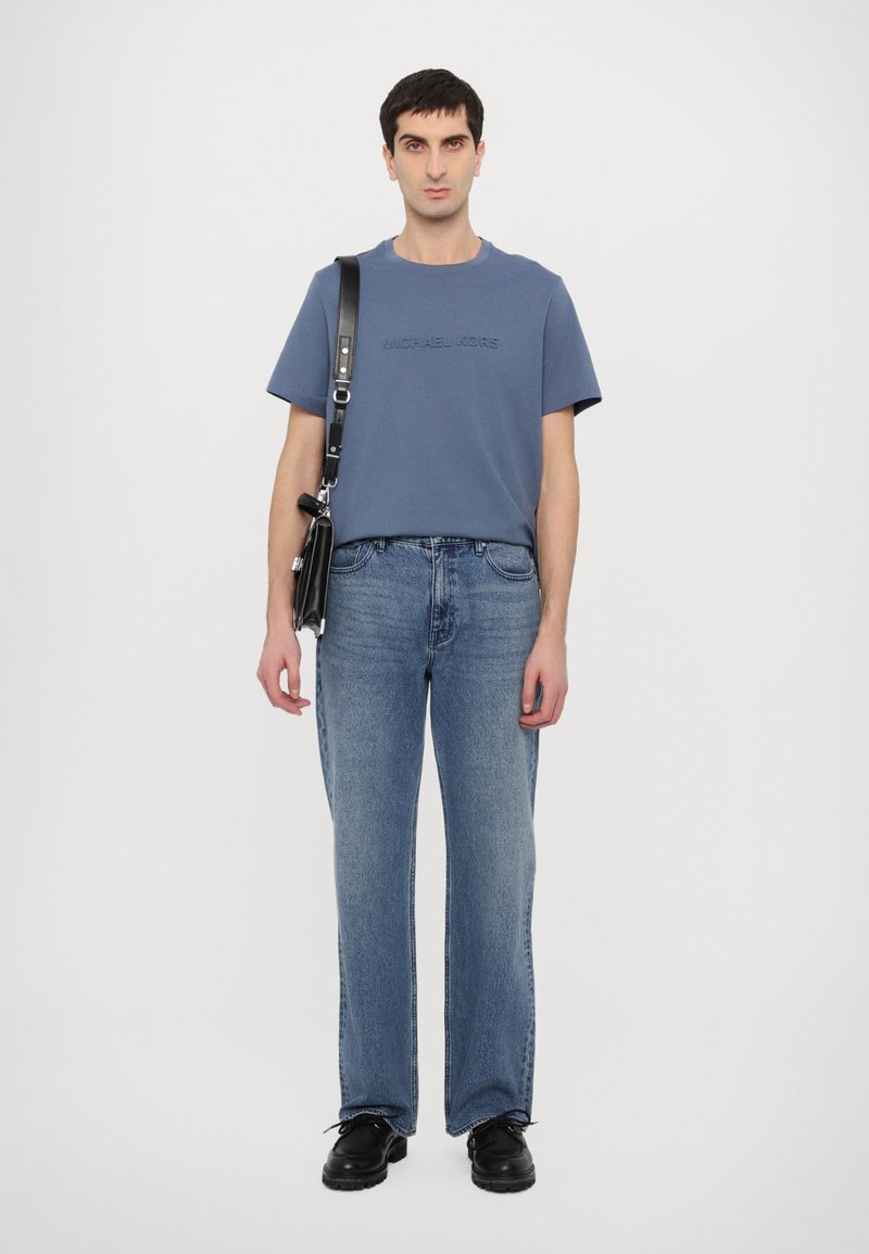 Blaues Baumwoll-T-Shirt mit geprägt "MICHAEL KORS" Logo, kombiniert mit hellblauen Jeans und schwarzer Schultertasche. Die Schuhe sind schwarz und klobig.