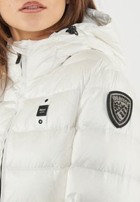 Giacca a vento bianca con finitura lucida, dotata di chiusura con zip, cappuccio regolabile e un badge nero con strass sul lato sinistro del petto.