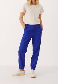 Blå byxor med elastisk midja och sidofickor, kombinerade med en beige t-shirt med kort ärm och vita sneakers med gula sulor.