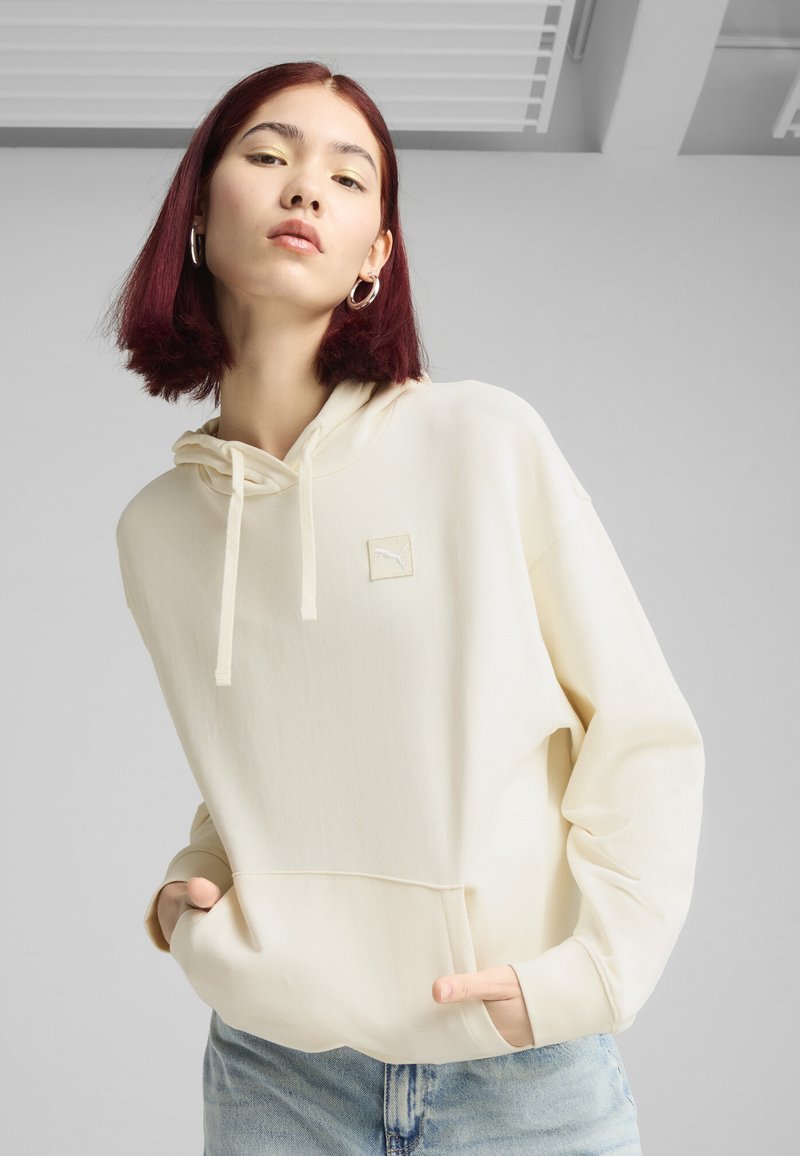 Sudadera color crema con una textura suave, cordones ajustables y un pequeño logo bordado en el pecho. Gran bolsillo frontal.