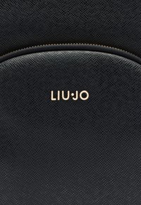 Melna teksturēta soma ar liektu formu, priekšpusē zelta krāsas "LIU•JO" logotips un zelta rāvējslēdzēja aizdare.