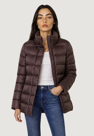 Donna che indossa una giacca imbottita trapuntata marrone scuro sopra una maglietta bianca e jeans blu, in piedi con una mano in tasca.