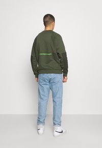 Green sweatshirt with dark sleeves, featuring the text "CONQUER YOUR FEAR OF HEIGHTS" in bright green. Paired with light blue jeans and white sneakers.