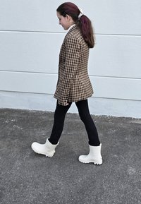 Bruine geblokte blazer, zwarte skinny broek en witte rubberen enkellaarsjes met gestructureerde zolen. Lopend tegen een lichtgrijze muur.