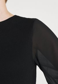 Svart stickad topp med genomskinlig svart tyg på ärmarna. Rund halsringning, figursydd kropp, strukturerad stickning på huvudplagget, slät ytfinish på ärmarna.