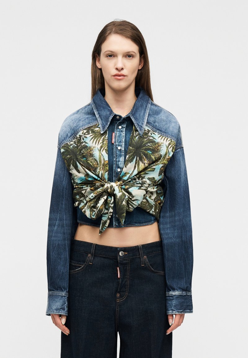 Γυναίκα που φορά cropped τζιν μπουφάν με μακριά μανίκια και ύφασμα με τροπικό σχέδιο δεμένο με κόμπο στο στήθος, συνδυασμένο με σκούρα τζιν παντελόνια.