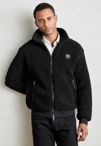 Giacca in pile nera con colletto alto, zip frontale e tasche laterali. Presenta un tessuto testurizzato e una toppa con logo sul petto.