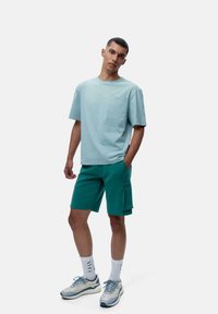 Šviesiai mėlynas medvilninis marškinėliai su kišenėle ir tekstiniu akcentu, derinami su žaliomis šortais, turinčiais šoninės kišenės ir priderintais rankogaliais. Pilki sportbačiai užbaigia įvaizdį.