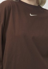 Braunes Baumwoll-T-Shirt mit Rundhalsausschnitt. Verfügt über ein kleines weißes Nike-Logo auf der Brust. Kurze Ärmel und entspannter Schnitt. Weicher Stoff.