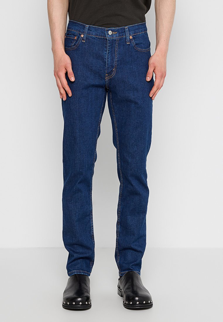 Jean en denim bleu foncé avec une coupe droite, cinq poches, des surpiqures orange et une fermeture à bouton à la taille.