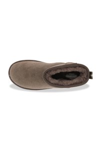Zapatilla de ante marrón con un interior suave y aterciopelado. Diseño sin cordones con bordes cosidos y una pequeña asa en el talón. Suela plana.
