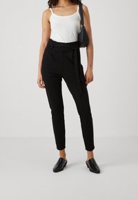 Žena stojí v černém topu a kalhotách, drží malou černou kabelku a předvádí ležérní, přesto stylový outfit na jednolitém pozadí.